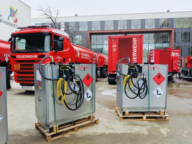 Mehrere rote Feuerwehrfahrzeuge und silberne Gefahrstoffbehälter mit Warnsymbolen stehen auf einem Hof vor einem modernen Gebäude.