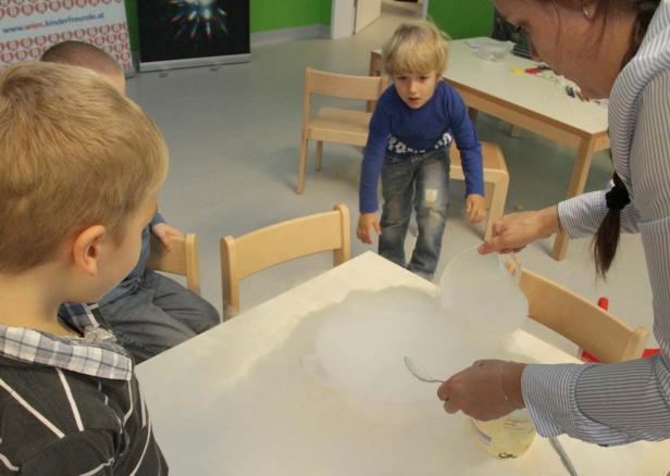 Kinder beobachten, wie eine Frau Trockeneis in einen Behälter gibt, wodurch Rauch entsteht.