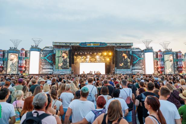 Viele Menschen stehen vor einer großen, bunt beleuchteten Konzertbühne mit mehreren Bildschirmen und genießen die Musik.