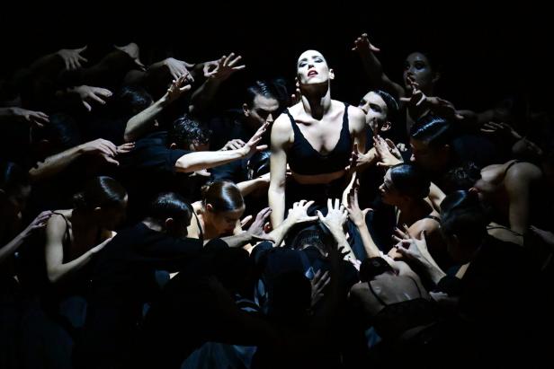 Eine Frau in schwarzer Kleidung steht im Mittelpunkt, umgeben von vielen ausgestreckten Armen anderer Tänzer.