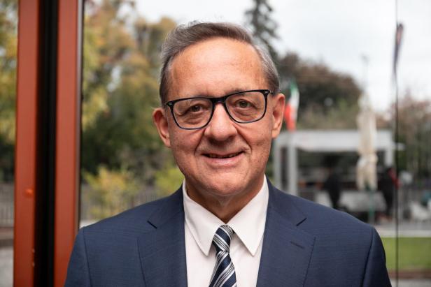 Ein älterer Mann mit Brille, Anzug und Krawatte steht lächelnd vor einem unscharfen, grünen Hintergrund.