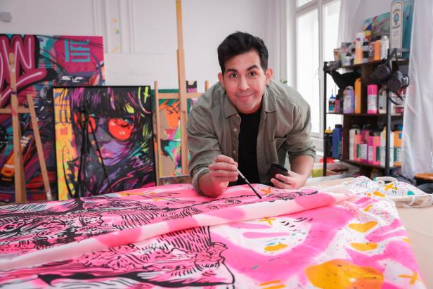 Ein Mann bemalt eine große, bunte Leinwand mit pinken und gelben Akzenten in einem Atelier mit Pop-Art-Gemälden.