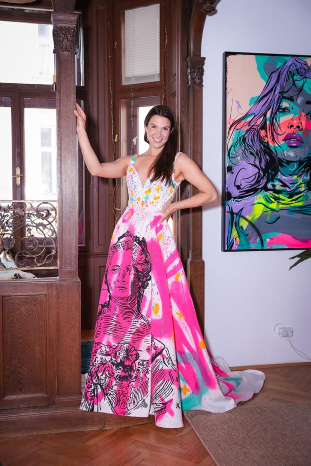 Eine Frau trägt ein langes, buntes Kleid mit auffälligem Porträtmotiv und steht lächelnd in einem stilvollen Raum.