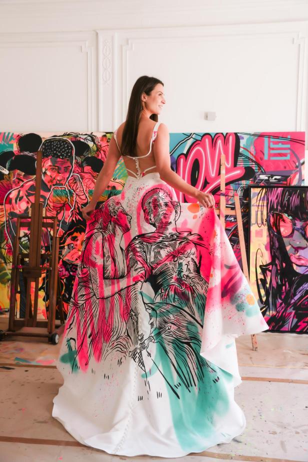 Frau in weißem Kleid mit buntem, handbemaltem Maria-Theresia-Motiv vor Pop-Art-Gemälden.