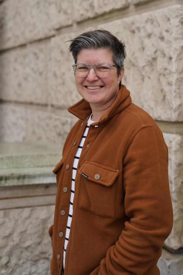 Person mit kurzer, grauer Frisur, Brille und brauner Jacke steht lächelnd vor einer hellen Steinwand.