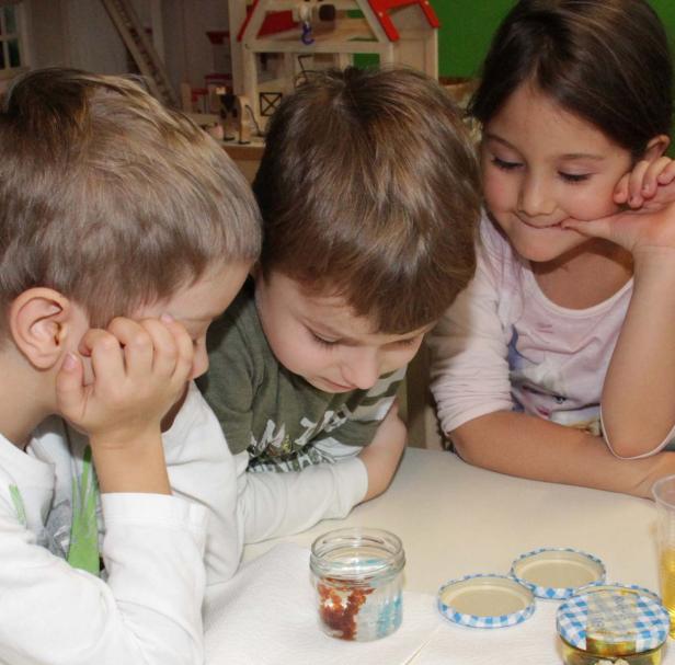 Drei Kinder betrachten ein Glas mit einer Flüssigkeit und Dekorationen darin.