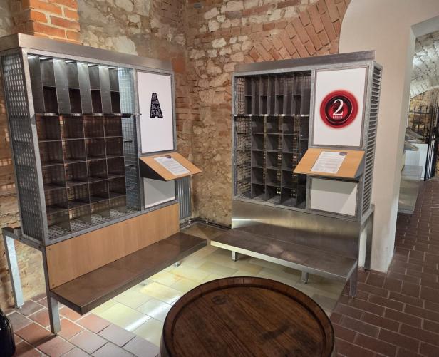Zwei Metallregale mit nummerierten Fächern und Infotafeln stehen vor einer Backsteinwand in einem historischen Raum.