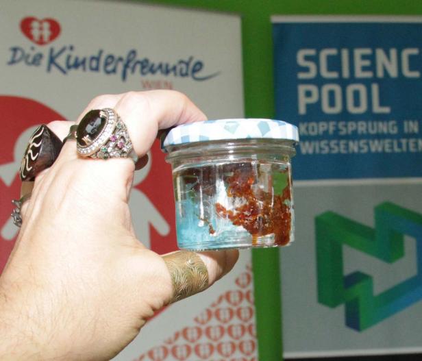 Eine Hand hält ein Glas mit einer braunen Substanz und blauer Flüssigkeit vor Bannern.