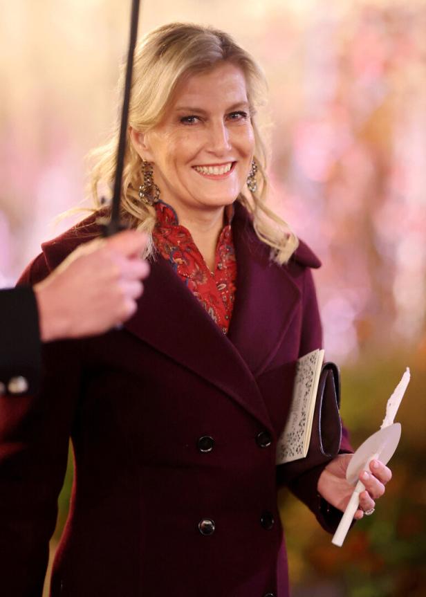 Eine lächelnde Herzogin Sophie mit blonden Haaren trägt einen burgunderroten Mantel und hält eine Kerze und eine Broschüre.