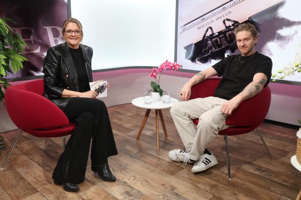 Zwei Personen sitzen in roten Sesseln in einem modernen Studio, zwischen ihnen steht ein kleiner Tisch mit Blumen und Wassergläsern.