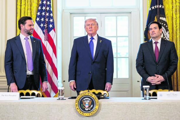 Donald Trump steht mit zwei Männern, Marco Rubio und The Vice President, vor US-Flaggen.