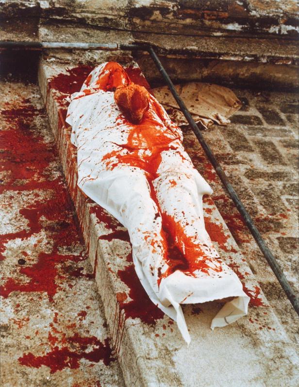 Eine in ein weißes Tuch gehüllte Figur liegt auf einem Steinsims, bedeckt mit roter Farbe.