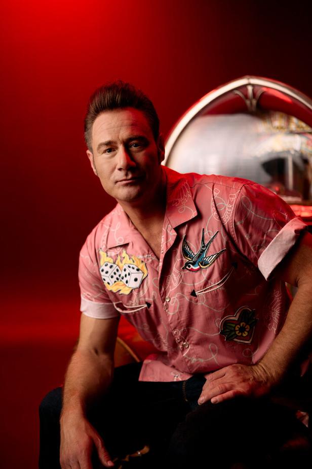 Ein Mann mit braunen Haaren trägt ein rosa Hemd mit bunten Motiven und sitzt vor einem rot beleuchteten Hintergrund.