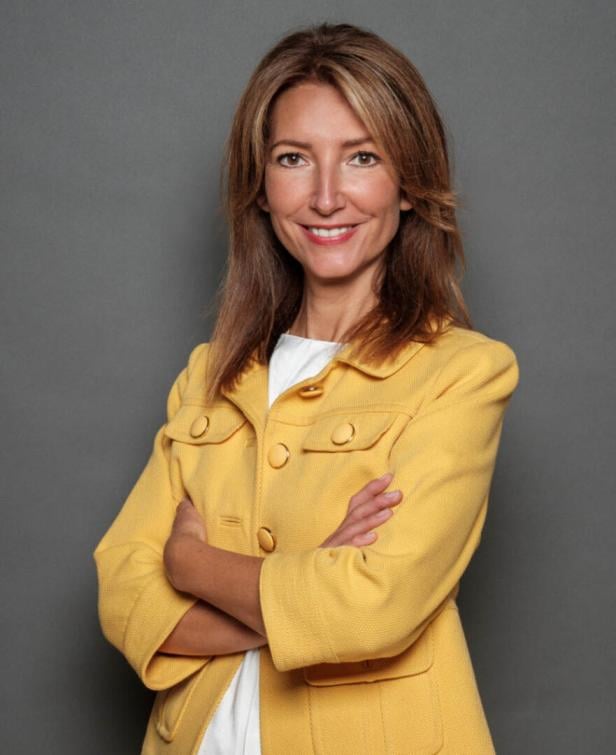 Frau mit langen braunen Haaren trägt ein gelbes Jackett und lächelt mit verschränkten Armen vor grauem Hintergrund.