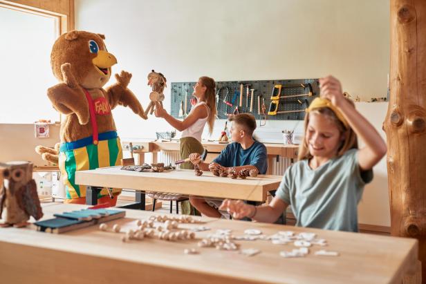 Kinder basteln mit Naturmaterialien, daneben steht ein Maskottchen im bunten Kostüm.