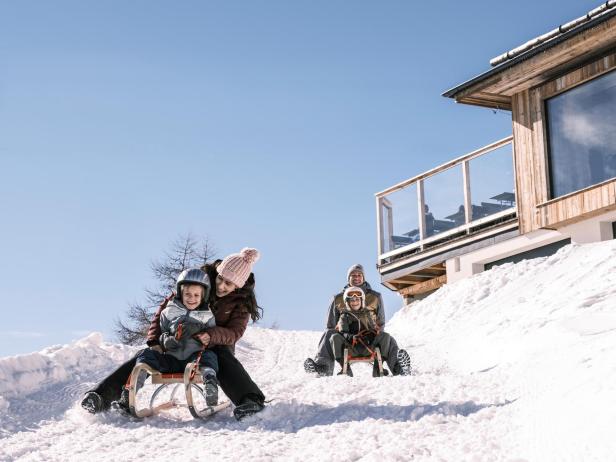 Zwei Familien rodeln lachend auf verschneiter Piste vor einer Berghütte im Sonnenschein.