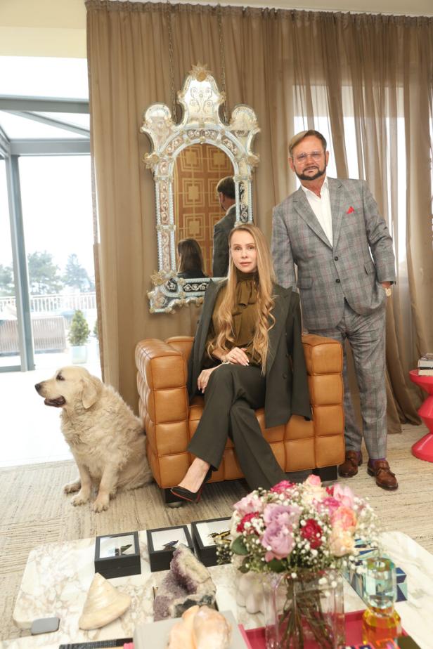 Ein Mann im Anzug steht neben einer Frau auf einem Sessel, ein Hund sitzt daneben, im Vordergrund ein Tisch mit Blumen und Dekoration.