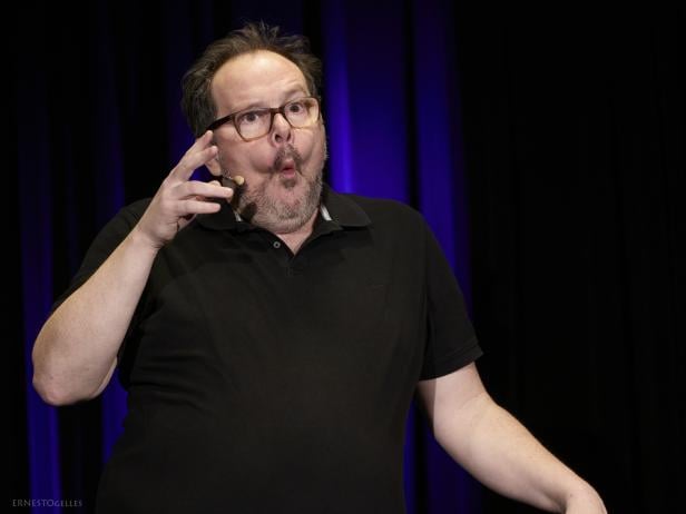 Ein Mann mit Brille und Mikrofon gestikuliert ausdrucksstark auf einer Bühne vor dunklem Hintergrund.