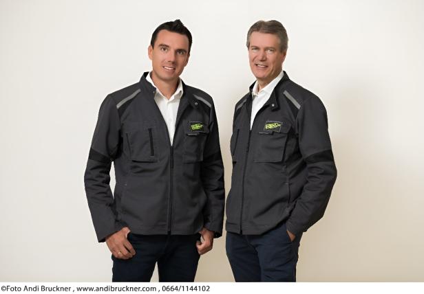 Zwei Männer in dunklen Arbeitsjacken mit Logo stehen nebeneinander und lächeln vor hellem Hintergrund.