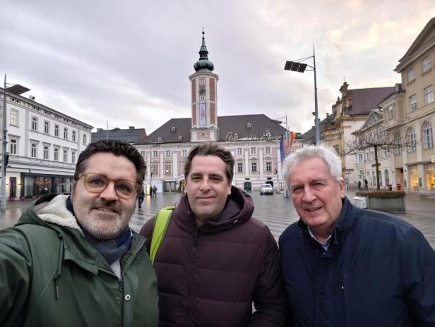 Drei Männer stehen auf einem Platz vor dem Rathaus von St. Pölten bei bewölktem Himmel.