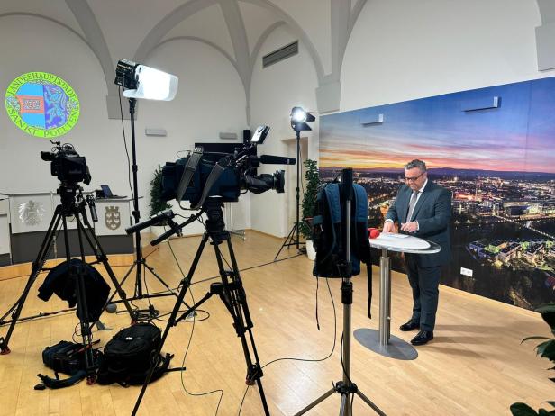 Ein Mann im Anzug steht an einem Stehtisch vor Kameras und Scheinwerfern in einem Studio.