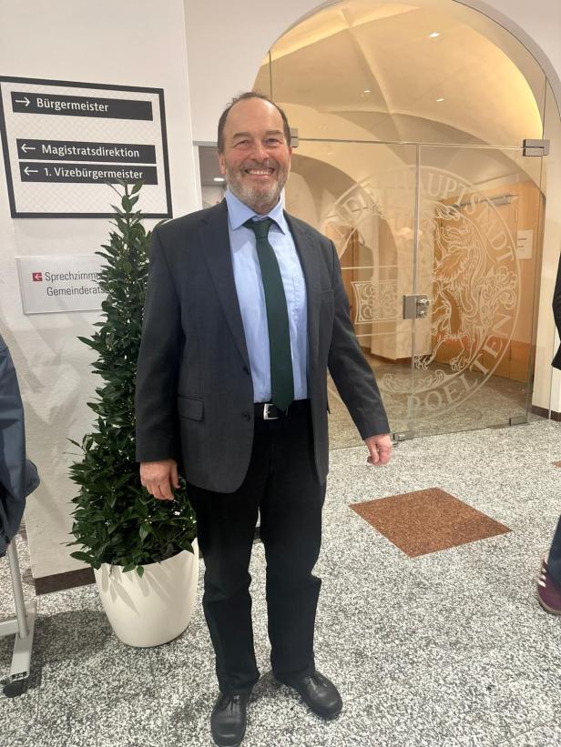 Ein Mann im Anzug steht lächelnd in einem Amtsgebäude neben Wegweisern.