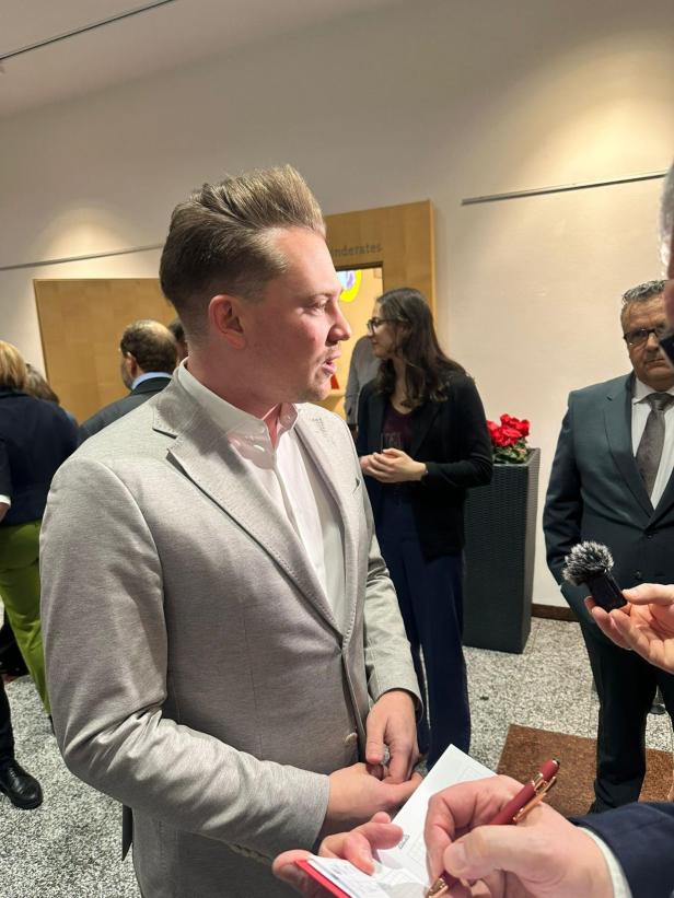 Florian Krumböck spricht bei einer Veranstaltung mit Journalisten, während im Hintergrund weitere Personen stehen.