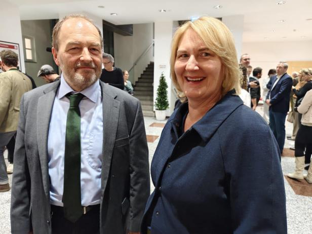 Zwei Personen stehen lächelnd in einem hellen Raum mit weiteren Menschen im Hintergrund.