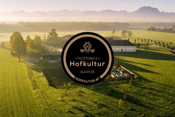 Das Logo von Hütthalers Hofkultur steht vor einem Bauernhof in ländlicher Umgebung und symbolisiert das Tierwohl-Projekt von XXXLutz für nachhaltige Lebensmittelqualität.