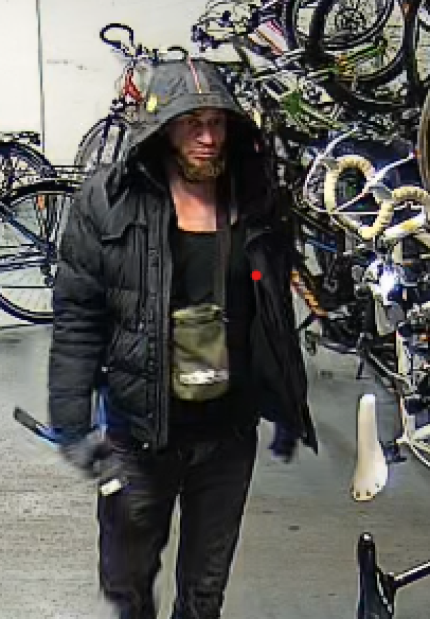 Eine Person mit dunkler Kapuzenjacke und Umhängetasche steht vor mehreren abgestellten Fahrrädern.