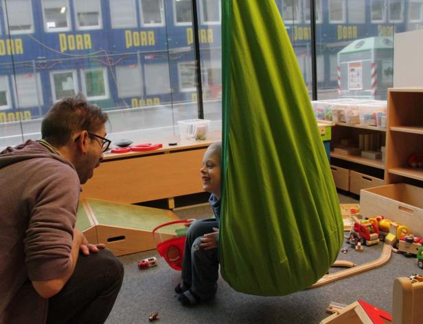 Ein Mann schaut einem Jungen zu, der in einer grünen Hängematte in einem Spielzimmer sitzt.