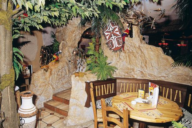 Ein Restaurantbereich ist mit Dschungelmotiven, Pflanzen, Felsen und einem Holztisch mit Stühlen dekoriert.
