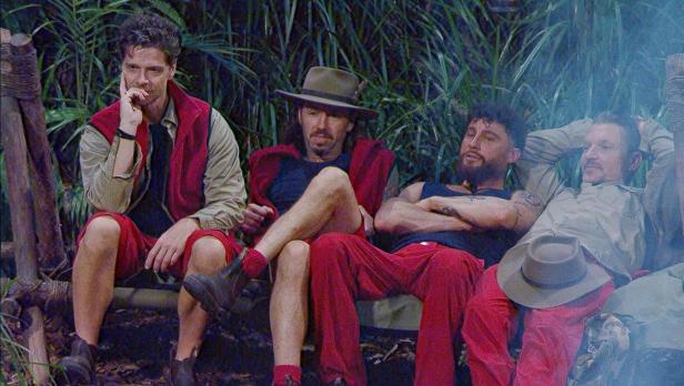Vier Männer in roter und beiger Kleidung sitzen und liegen entspannt auf einer Bank im Dschungel, umgeben von Pflanzen.