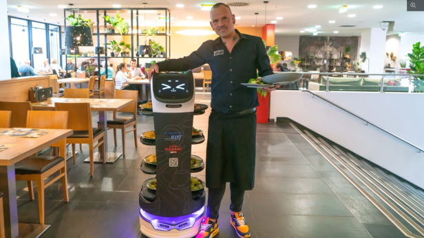 Ein Kellner steht neben einem Servierroboter mit mehreren Tellern Essen in einem modernen Restaurant.