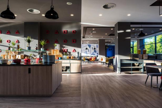 Modernes Hotelrestaurant mit Buffetbereich, bunten Stühlen und großen Fenstern mit Tageslicht.