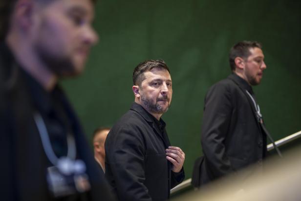 Wolodymyr Selenskyj in dunkler Kleidung, umgeben von weiteren Personen, vor einem grünen Hintergrund.