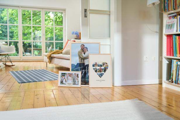 Gerahmte Fotocollagen und Paarfotos stehen auf dem Holzboden in einem hellen Wohnzimmer.