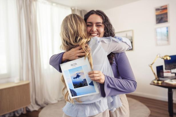 Zwei Frauen umarmen sich lächelnd, eine hält ein Fotobuch mit dem Titel „Ibiza“.