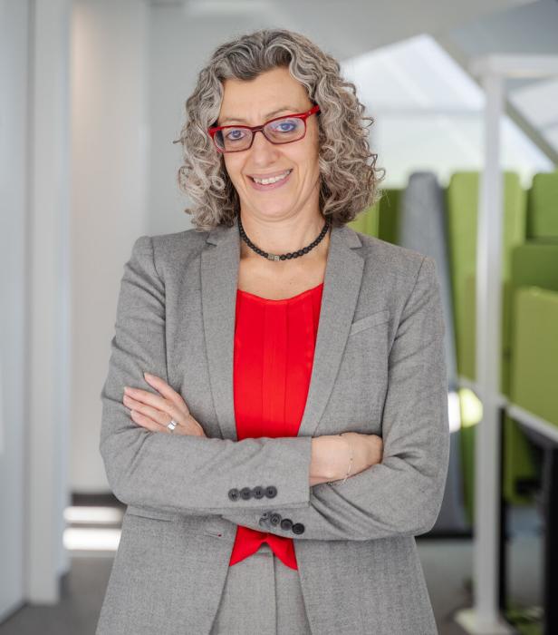 Frau mit lockigem grauem Haar, roter Brille und grauem Anzug steht mit verschränkten Armen und lächelt.