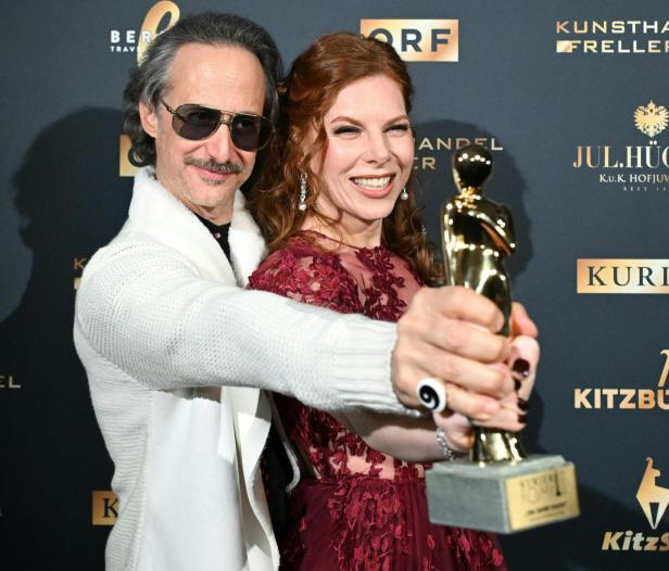 Ein Mann und eine Frau lächeln und halten gemeinsam eine goldene Trophäe vor einer Sponsorenwand.