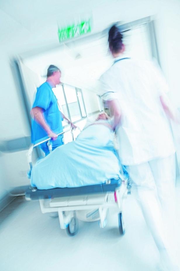 Zwei medizinische Fachkräfte schieben eine liegende Person auf einer Trage eilig durch einen Krankenhausflur.