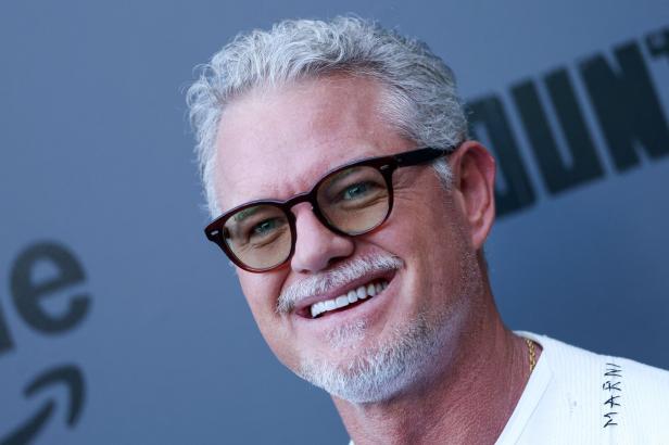 Ein lächelnder Eric Dane mit grauen Haaren und Brille vor einem grauen Hintergrund.