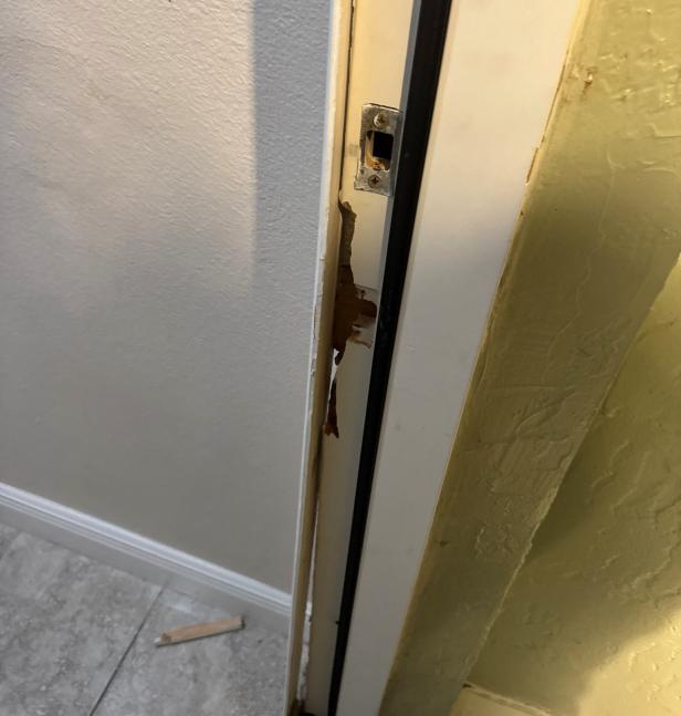 Beschädigter Türrahmen mit herausgebrochenem Holz und abgebrochenem Stück am Boden.