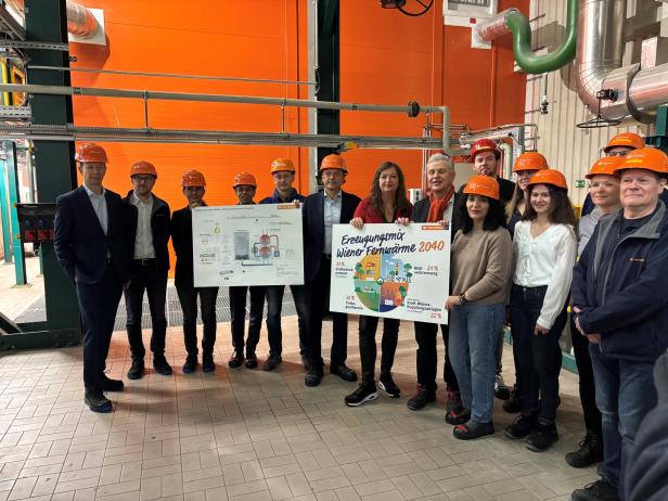 Eine Gruppe von Menschen mit orangefarbenen Helmen hält Schautafeln zu Fernwärme in einer Industriehalle.