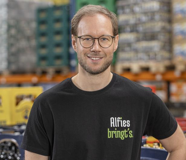 Ein Mann mit Brille und schwarzem T-Shirt mit der Aufschrift „Alfies bringt’s“ steht in einem Lager mit Getränkekisten.