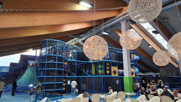 Ein Indoor-Spielplatz mit Klettergerüsten, Rutschen und sitzenden Erwachsenen unter dekorativen Lampen.
