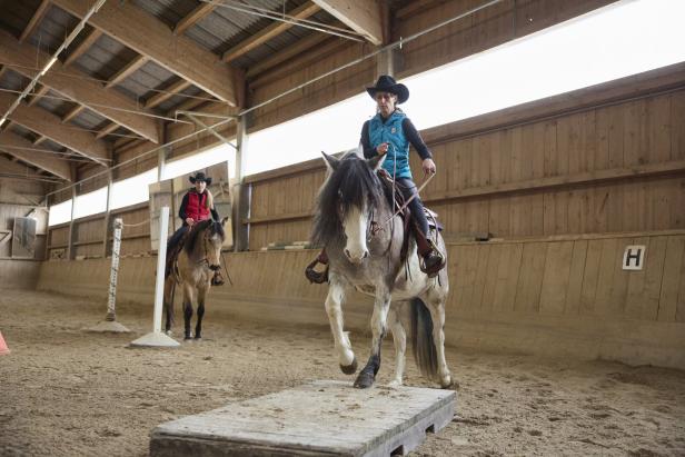 In einer Reithalle reitet eine Frau mit Cowboyhut ein Schimmelpferd über eine Holzplattform.