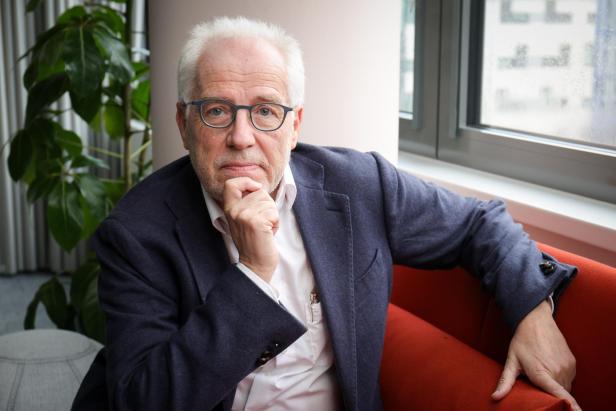 Ein älterer Mann mit Brille und weißem Haar sitzt nachdenklich auf einem roten Sofa neben einem Fenster und einer grünen Pflanze.