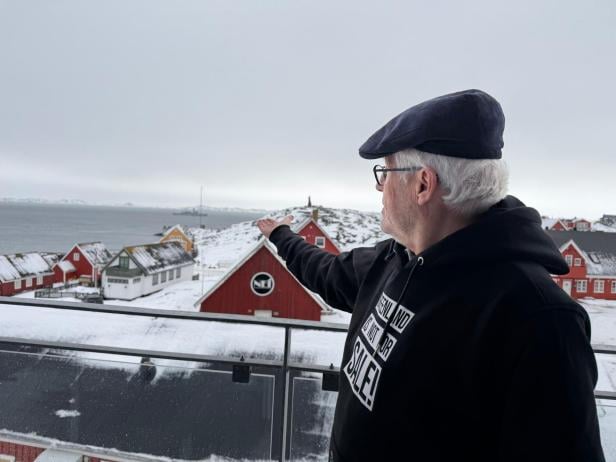 Ein Mann mit Mütze und Pullover ("Grönland steht nicht zum Verkauf") zeigt auf eine verschneite Küstenstadt.