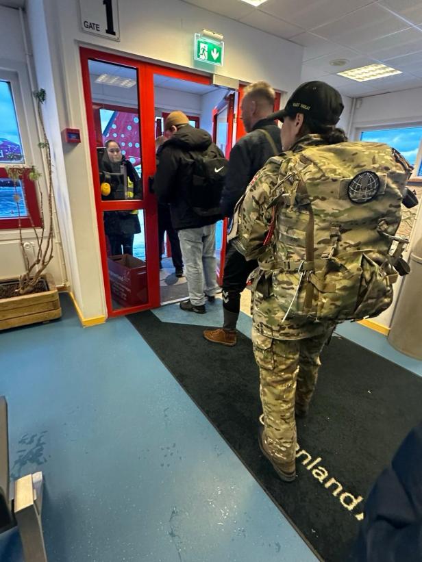 Mehrere Personen stehen in einer Schlange an einem Gate, darunter eine Person in Militärkleidung mit großem Rucksack.